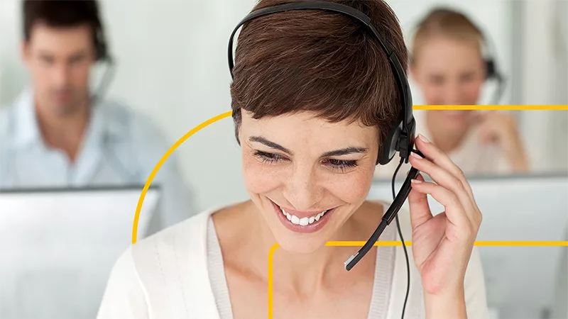 Foto einer lächelnden Frau mit einem HEadset auf dem Kopf, die in einem Call Center ein Telefonat entgegen nimmt