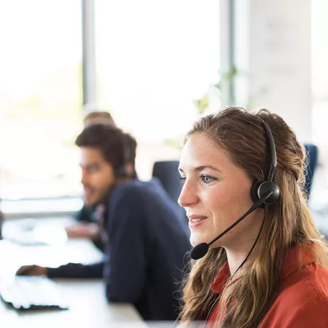 Foto einer Frau mit Headset, die in einem Großraumbüro sitzt und für Kundensupport zuständig ist